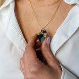 A person wearing a white shirt holds a unique jewelry piece—a gold anatomical heart necklace with a crown-shaped pendant. Their nails are painted a pale pink color, adding elegance to the handcrafted jewelry design.
