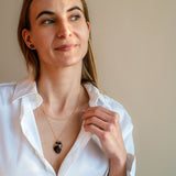 A woman with long light brown hair wears a white button-up shirt and a gold anatomical heart necklace, looking slightly to the side with a soft smile against a plain beige background.