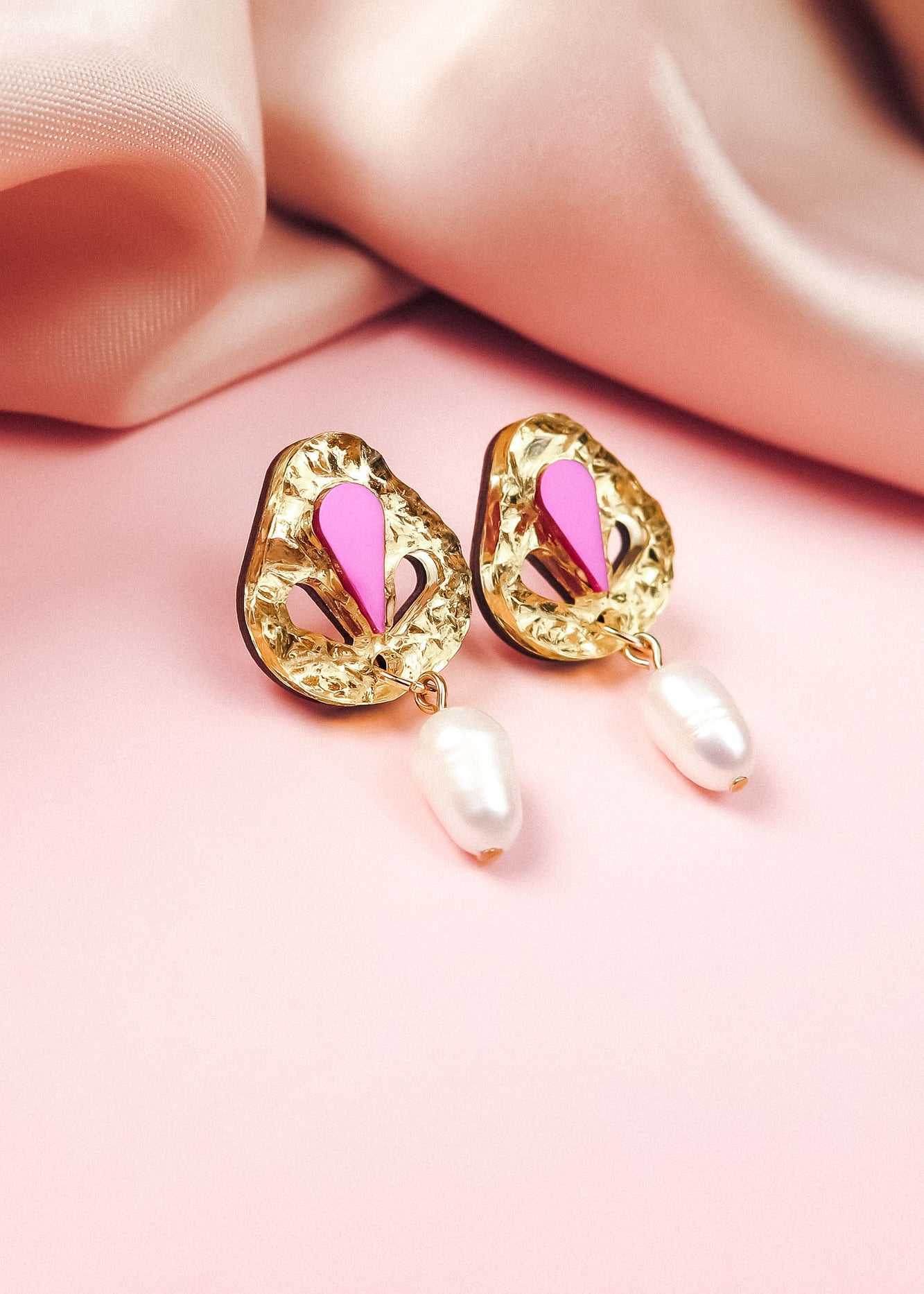 Handcrafted gold drop earrings with textured, abstract shapes, featuring pink and magenta accents and a dangling white pearl—perfect for minimalist fashion—displayed on a soft pink surface with silky pink fabric in the background.