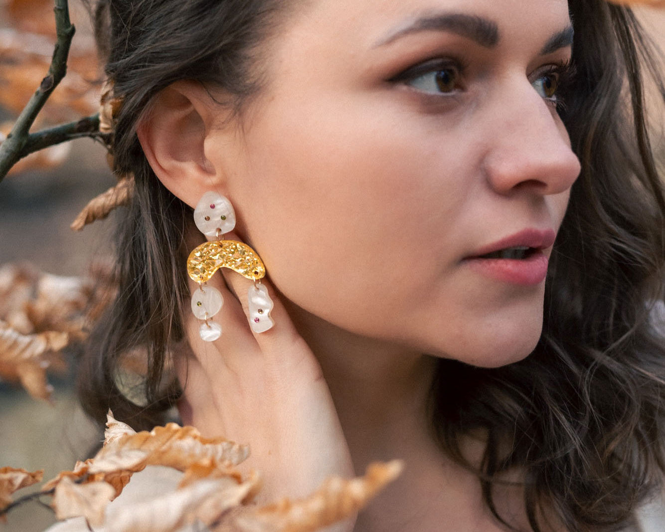 A woman with wavy brown hair wearing a cream top and boho chic dangle earrings stands among branches with dry, light brown leaves, gazing thoughtfully to the right.