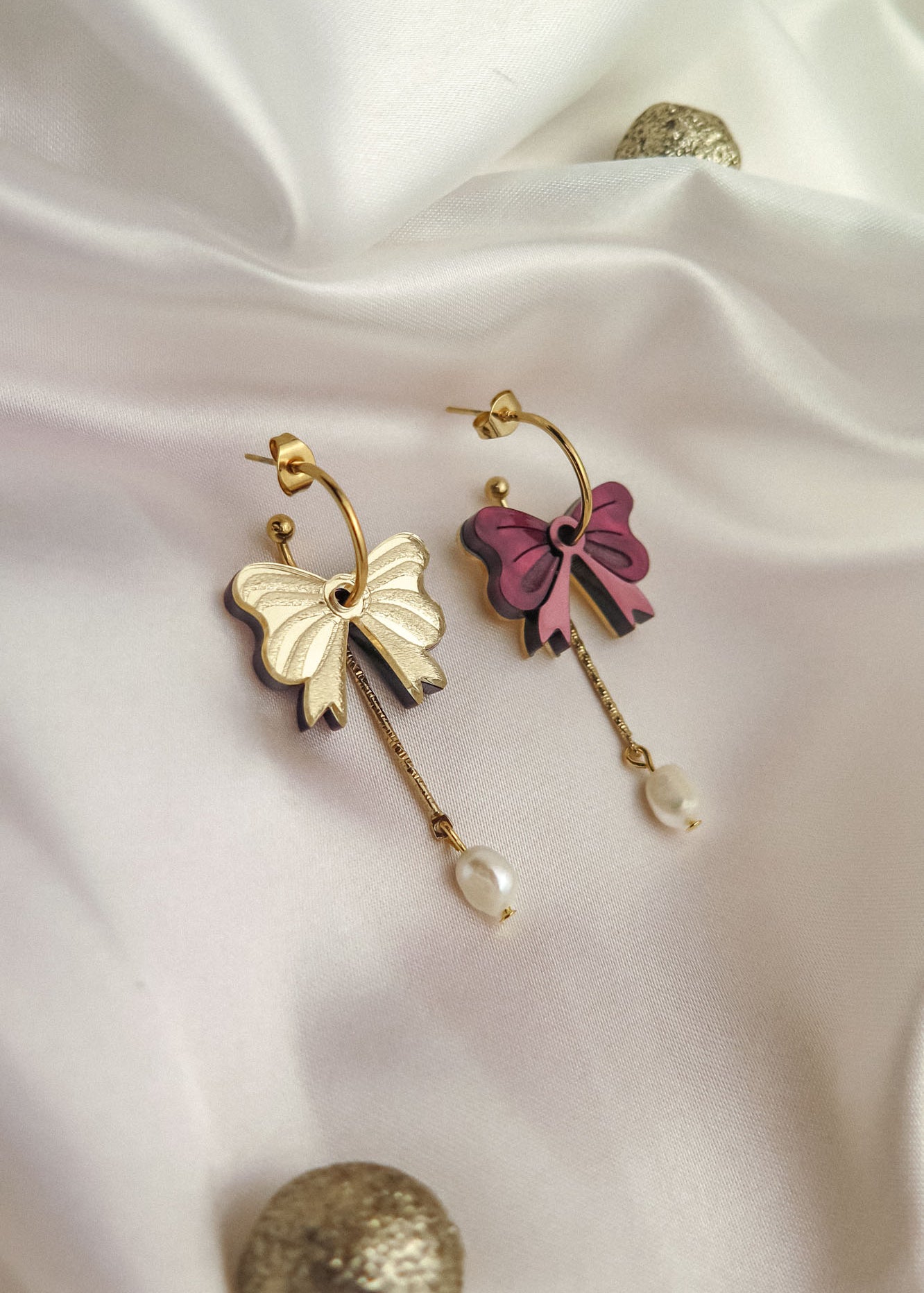 Two gold-plated hoop earrings with bow-shaped charms—one cream and one pink—each with a dangling gold chain and a freshwater pearl, displayed on white fabric with decorative gold spheres.