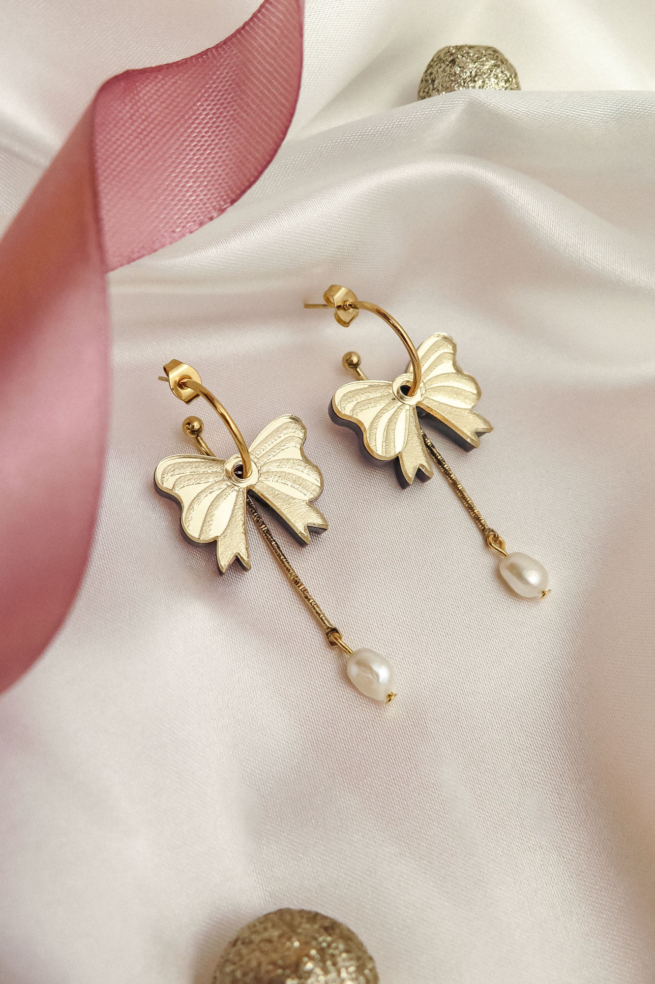 A pair of gold-plated earrings shaped like bows with dangling freshwater pearl accents, displayed on white satin fabric with a pink ribbon and gold decorative balls nearby.