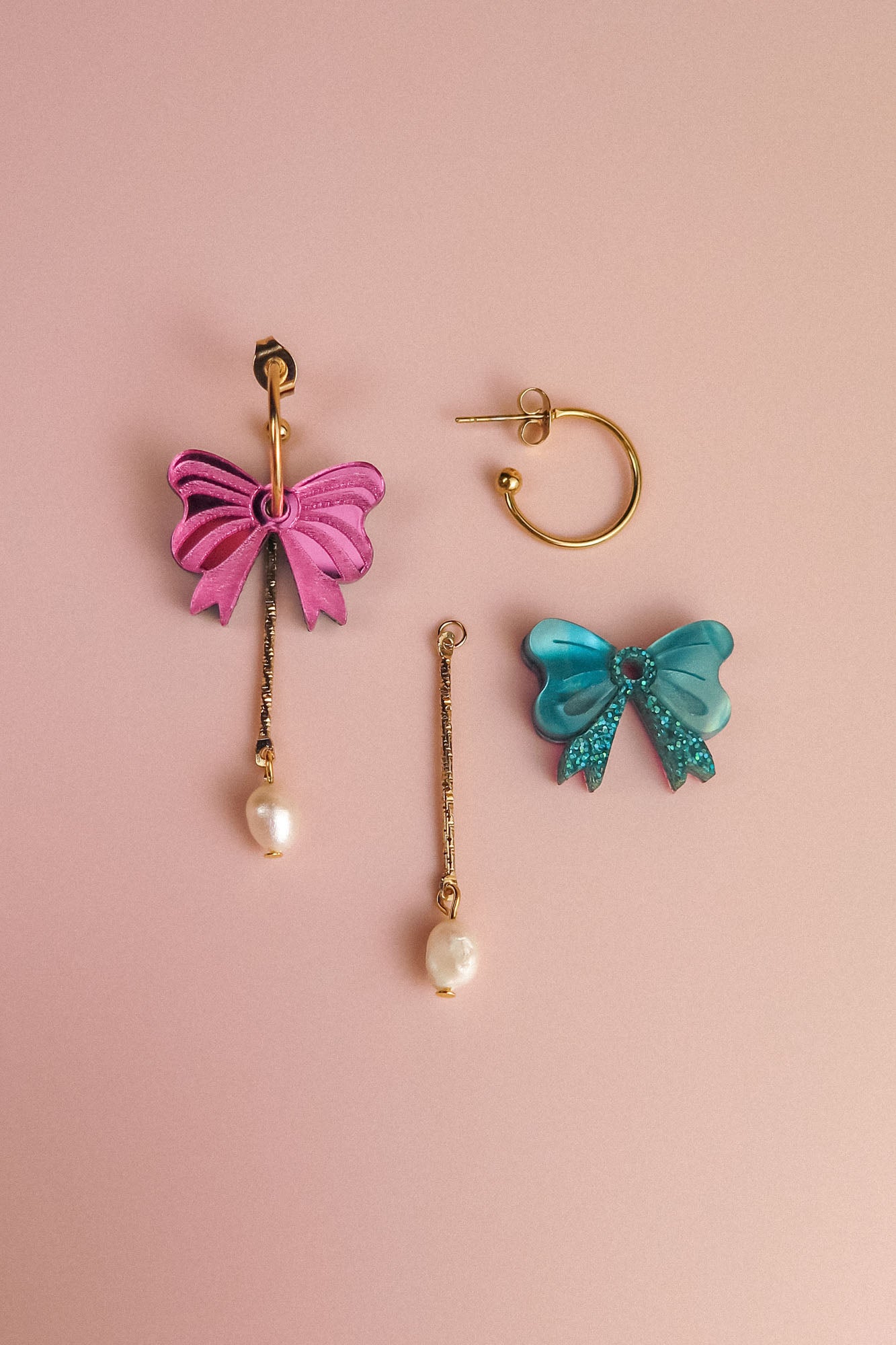 Three mismatched interchangeable dangle earrings on a pink surface: a gold-plated c-hoop, a dangling earring with a pink bow and freshwater pearl, and another with a blue bow, sequins, and pearl.