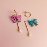 A pair of mismatched interchangeable dangle earrings on a pink background: one earring features a pink bow with a dangling freshwater pearl, while the other has a blue bow with a gold-plated c-hoop and bar, also adorned with a pearl.