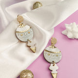 A pair of gold and white festive earrings with pearl details rest on white satin fabric, surrounded by a glittery gold ball and a white decorative bow on a lavender background.