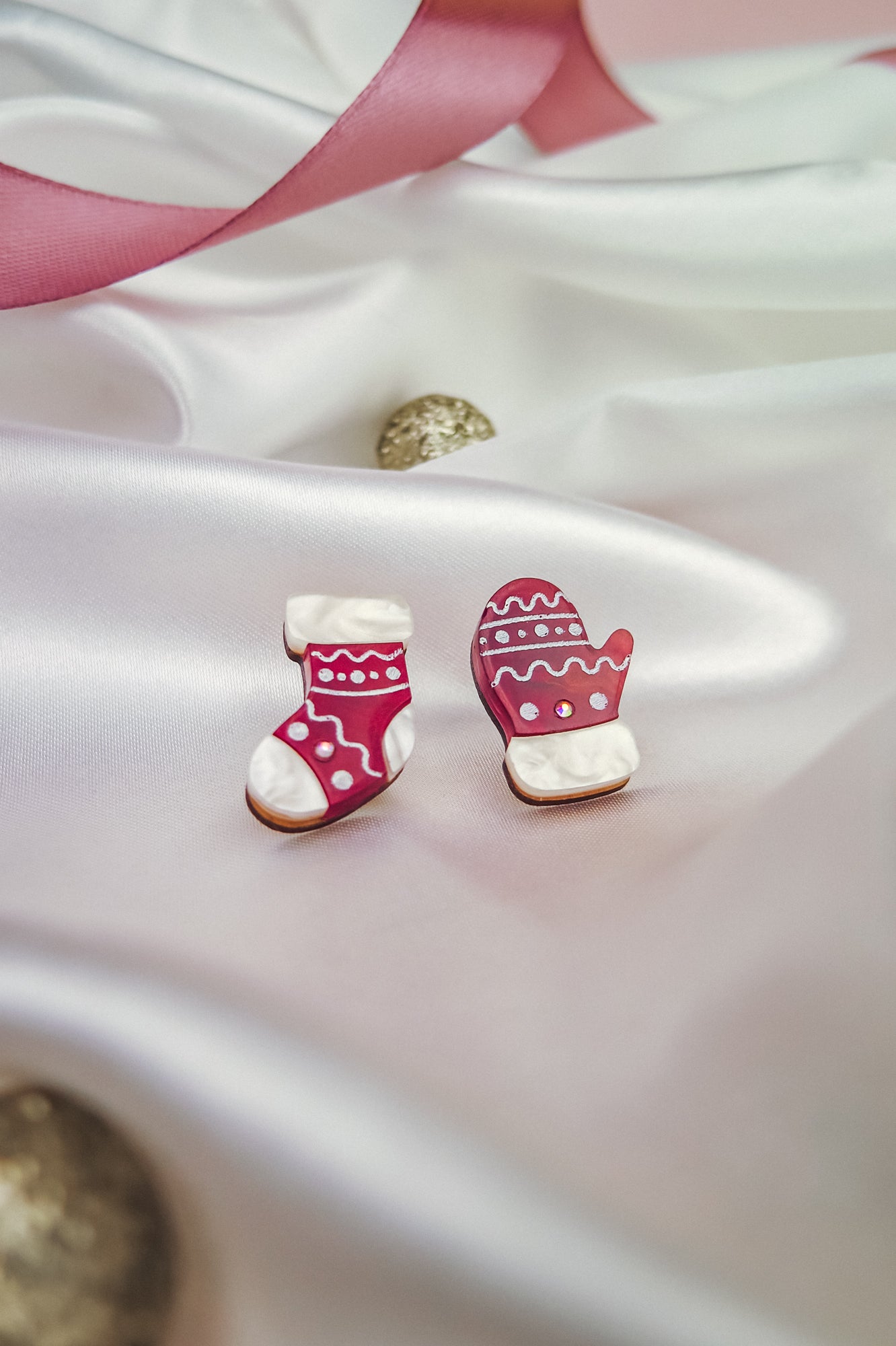 Red and white decorated cookies shaped like a Christmas stocking and mitten sit on silky white fabric, surrounded by festive accessories—a pink ribbon, gold ornaments, and charming Christmas Knitted Studs holiday earrings in the background.