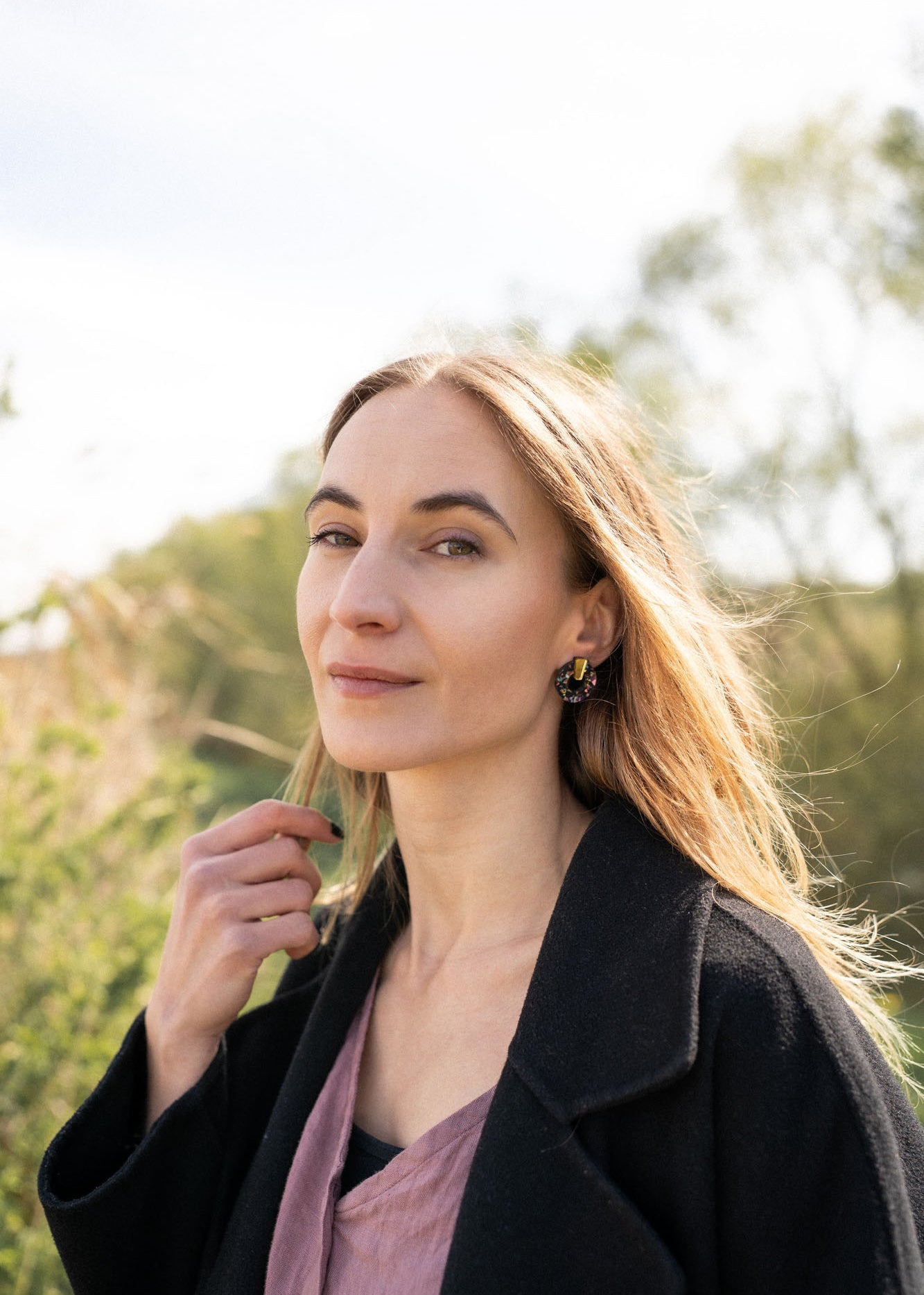 A woman with long, light brown hair stands outdoors, wearing a dark coat, mauve top, and minimalist circle stud earrings. She lightly touches her hair and looks calmly at the camera against green foliage and a bright, sunlit sky.