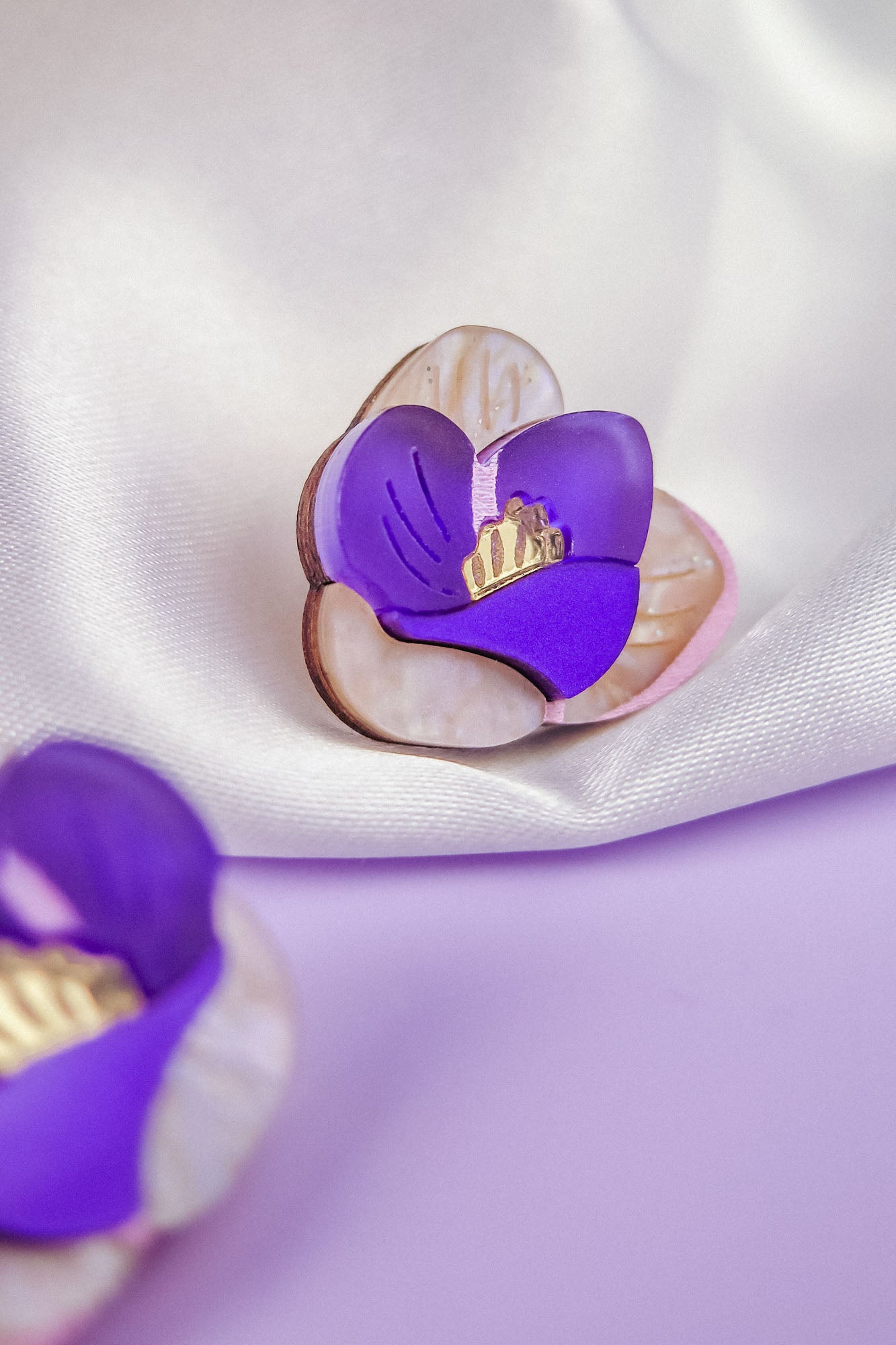 A decorative brooch shaped like a purple and cream hand-painted flower with gold details rests on silky white fabric. Another similar brooch is partially visible in the foreground.