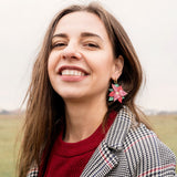A woman with long brown hair smiles outdoors, wearing a red sweater, a black-and-white checkered coat, and festive holiday earrings. The background is a grassy field under an overcast sky.