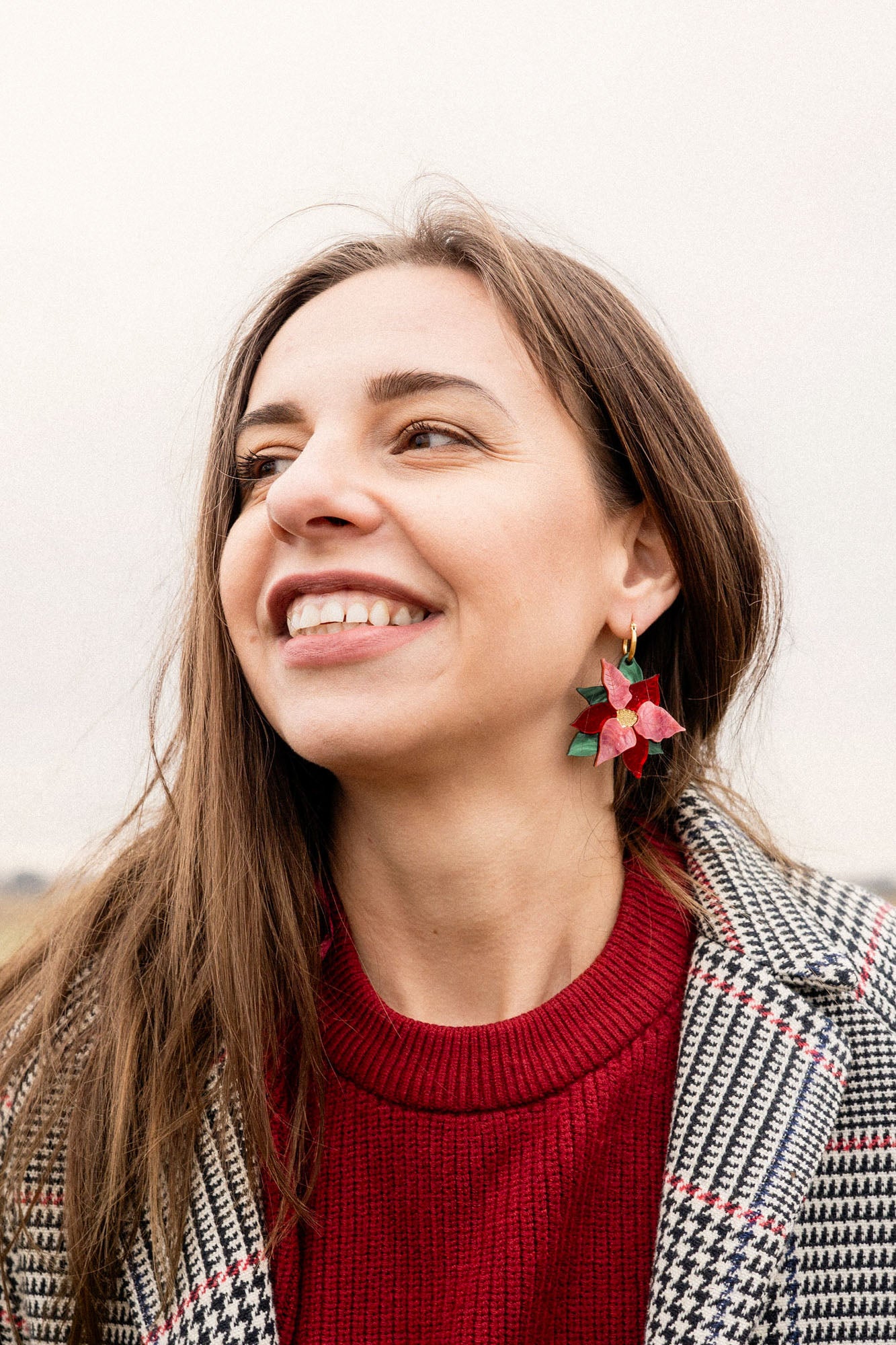 A woman with long brown hair smiles while wearing a red sweater, a checkered coat, and Poinsettia Statement Hoops, adding a touch of festive jewelry. The background is outdoors with a cloudy sky.
