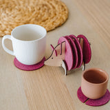 A white mug and a pink cup rest on purple felt coasters, next to a hedgehog-shaped coaster holder with more coasters on a light wooden table. A round woven placemat is in the background—a charming housewarming gift idea.