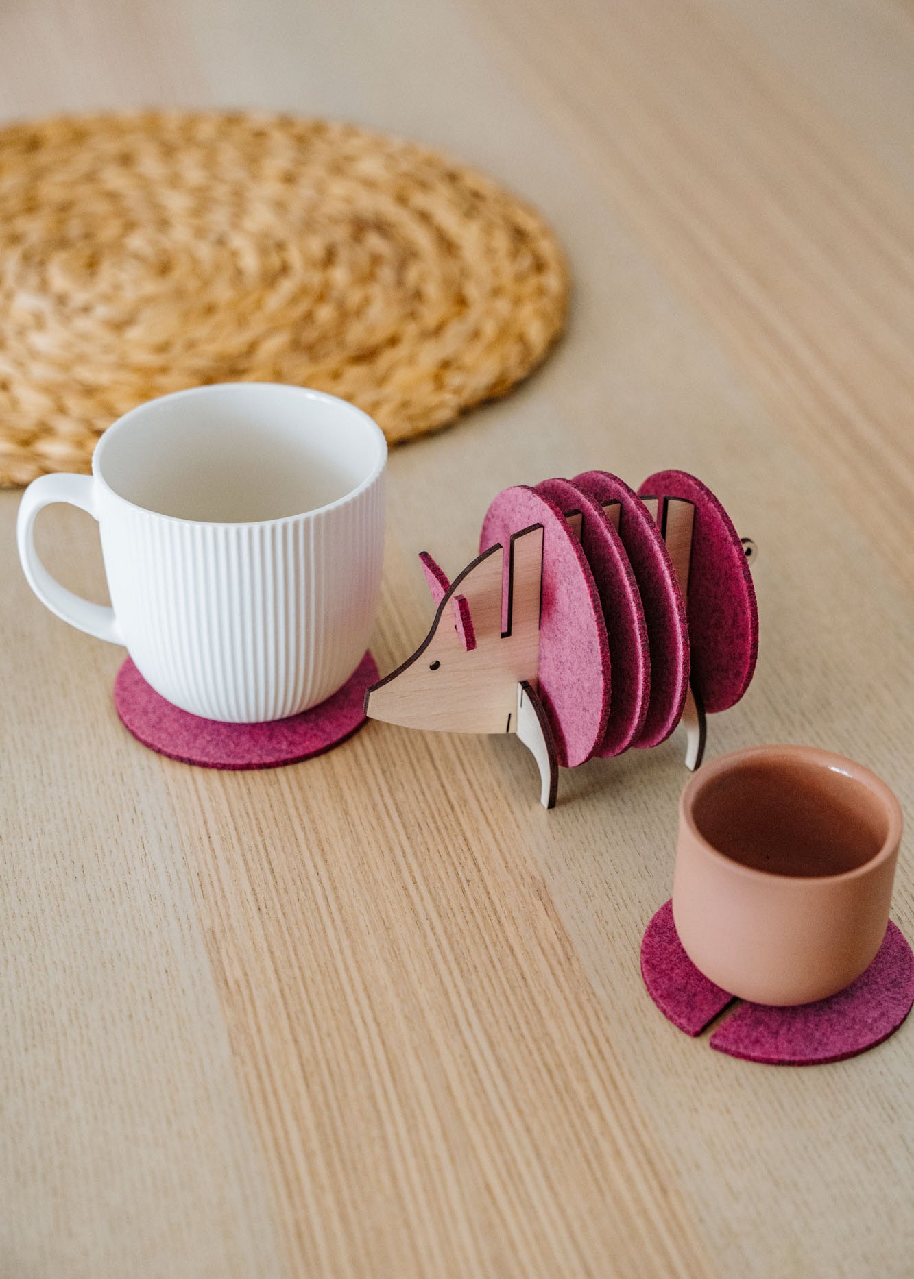 A white mug and a pink cup rest on purple felt coasters, next to a hedgehog-shaped coaster holder with more coasters on a light wooden table. A round woven placemat is in the background—a charming housewarming gift idea.
