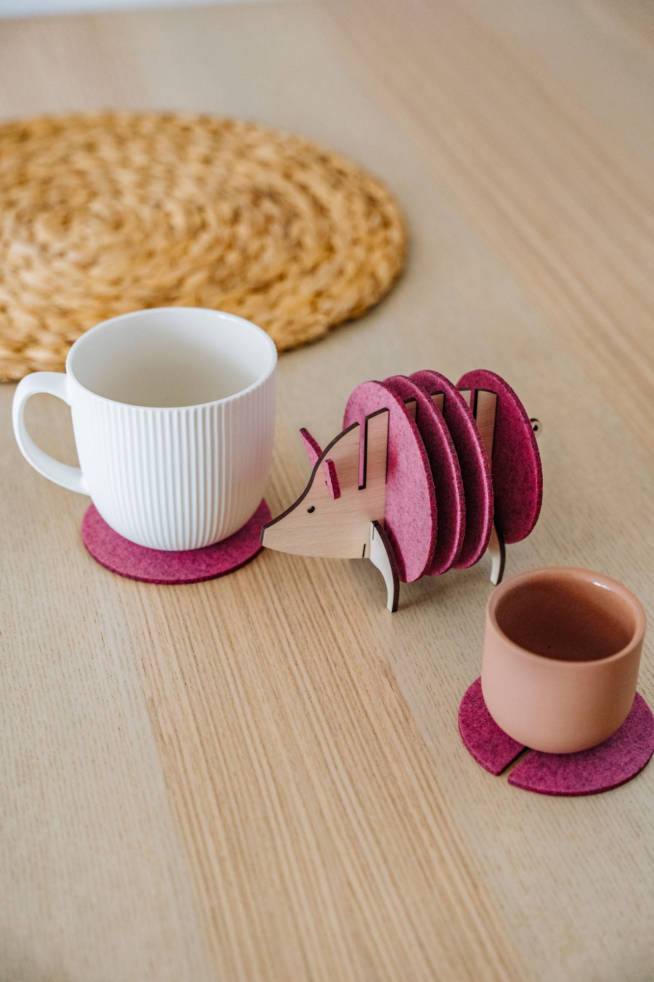 A white mug and a pink cup rest on purple felt coasters, next to a hedgehog-shaped coaster holder with more coasters on a light wooden table. A round woven placemat is in the background—a charming housewarming gift idea.