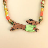 A colorful, handcrafted Dachshund necklace featuring a patterned cord and a pendant shaped like a brown dachshund in a green sweater, made from recycled materials and shown against a beige background.