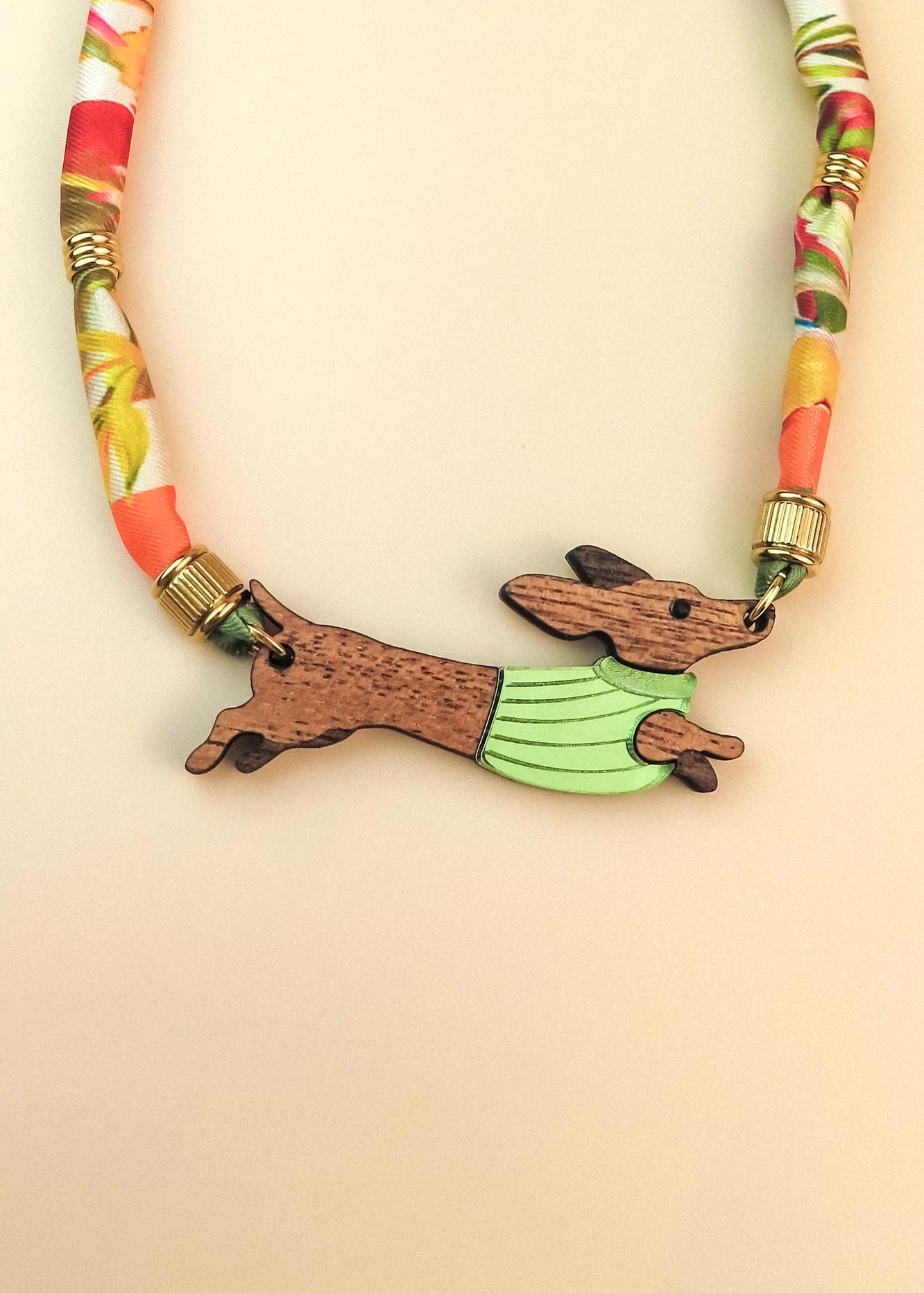 A colorful, handcrafted Dachshund necklace featuring a patterned cord and a pendant shaped like a brown dachshund in a green sweater, made from recycled materials and shown against a beige background.