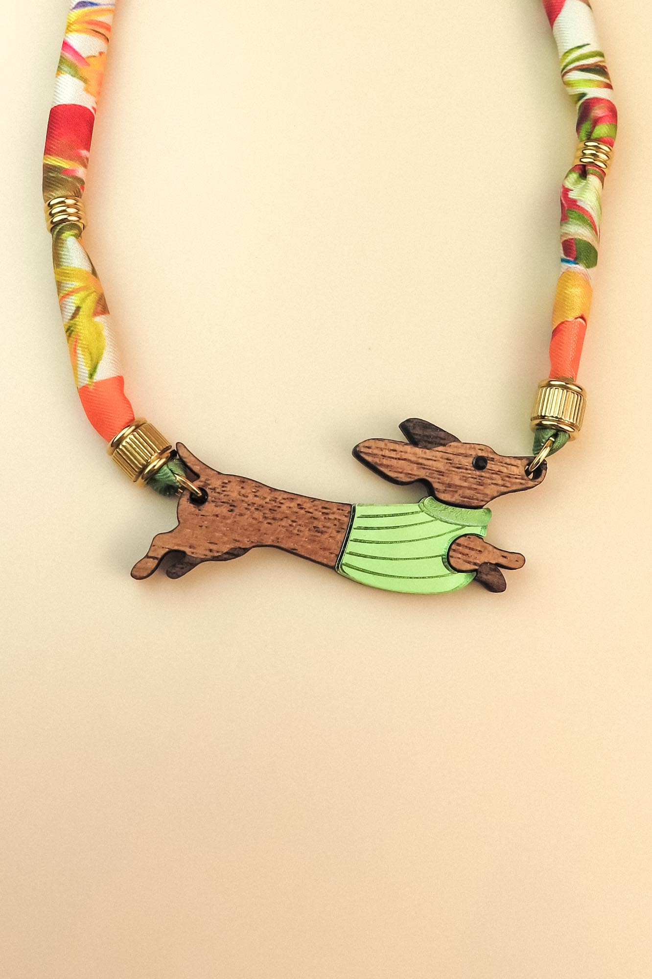A colorful, handcrafted Dachshund necklace featuring a patterned cord and a pendant shaped like a brown dachshund in a green sweater, made from recycled materials and shown against a beige background.