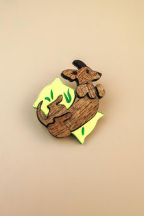 A playful brooch shaped like a dachshund lies on a plain beige background. With engraved details and bright yellow and green leaf-like accents, this wooden piece adds whimsy to any dog accessory collection.