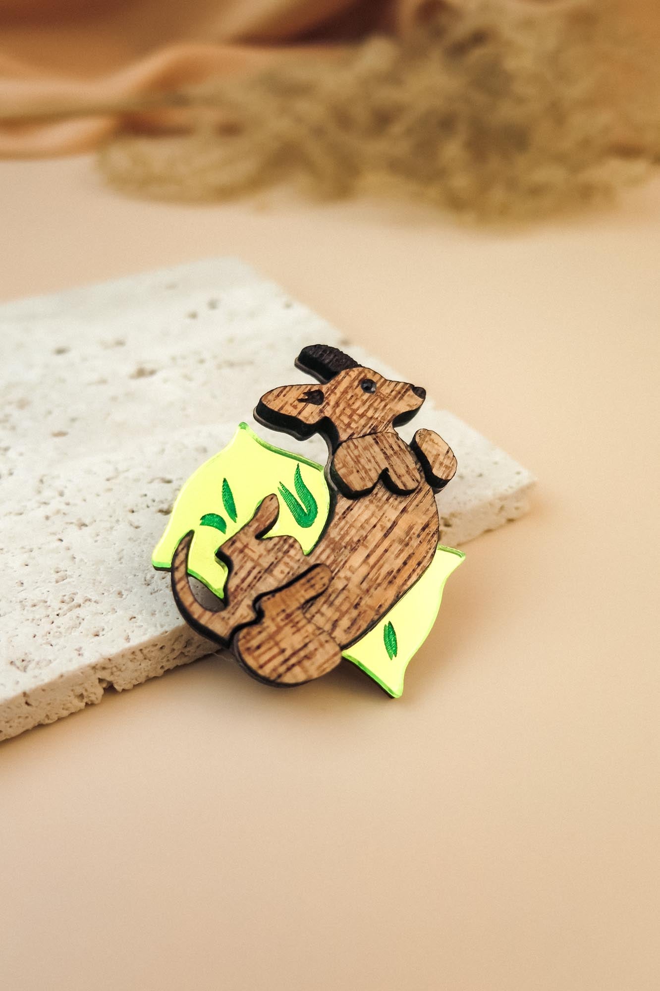 A playful brooch, this wooden pin shaped like a mouse lounging on a yellow wedge with green leaf accents rests on textured beige stone, making it a charming dog accessory or a unique Dachshund brooch for any outfit.