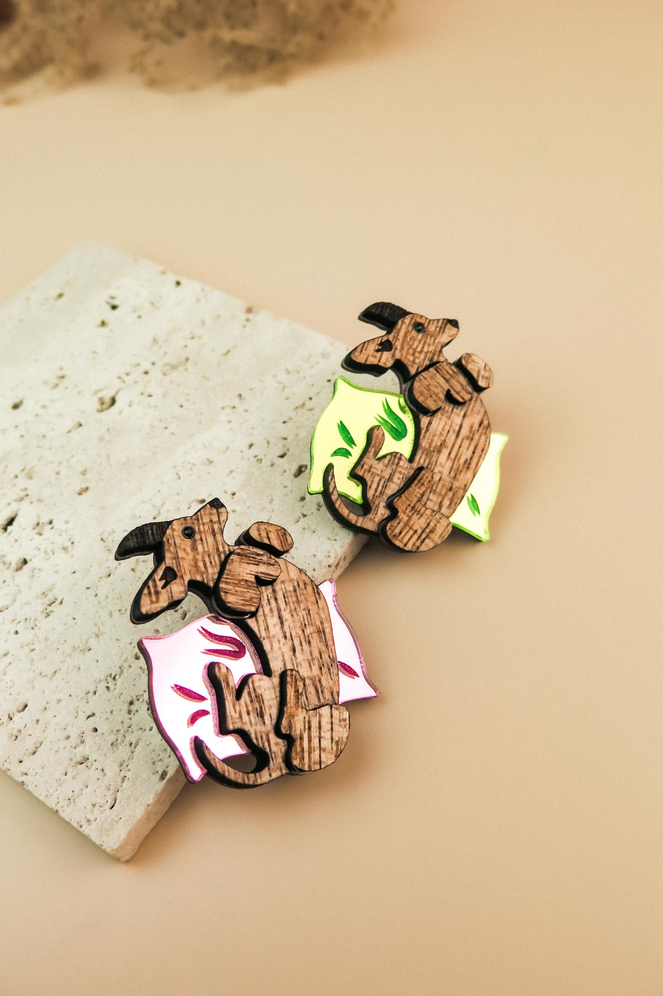 Two playful brooches shaped like reclining capybaras, each resting on a colorful pillow—one pink and one neon green—placed on a textured stone surface. A charming touch for any dog accessory collection or fans of unique brooches.
