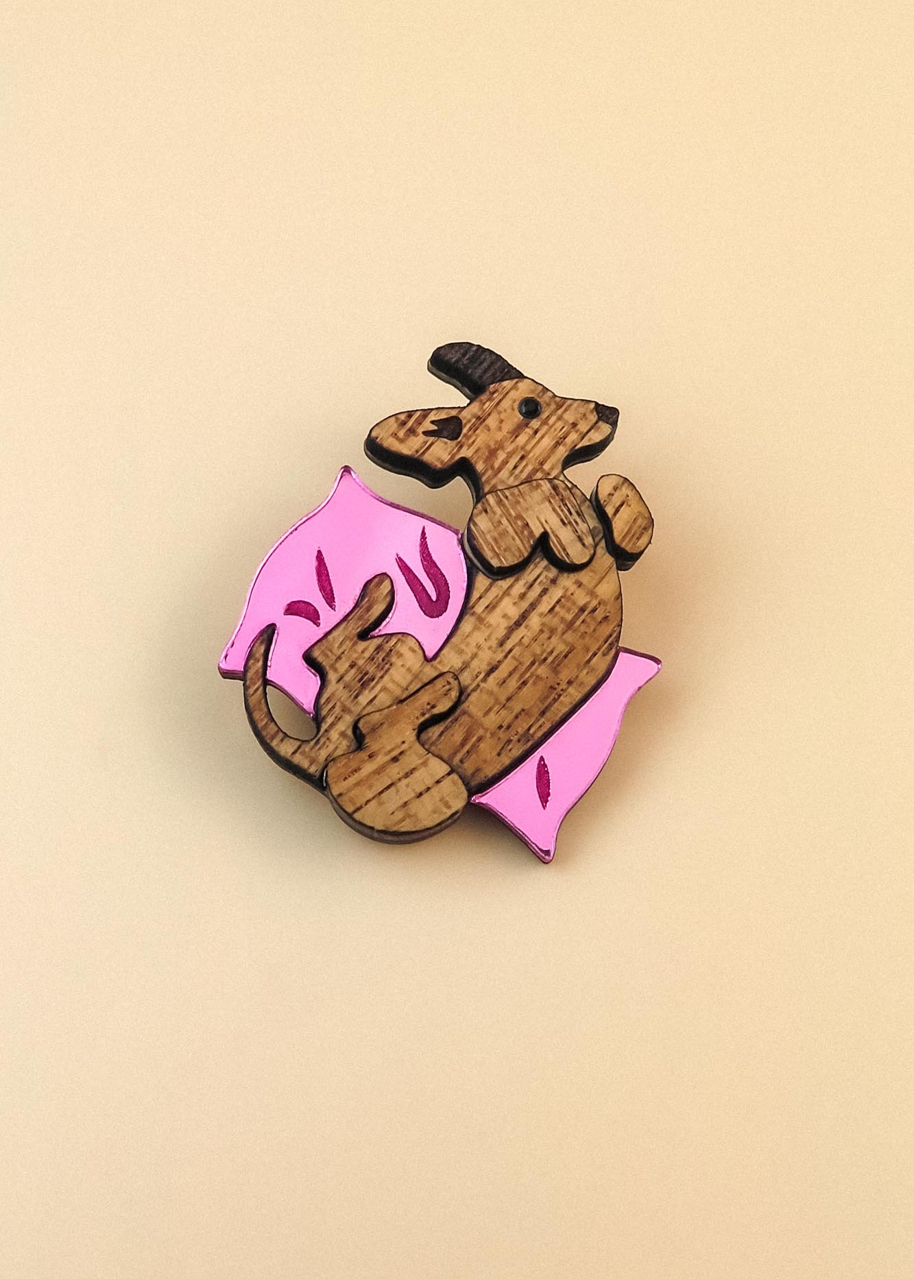 A wooden Dachshund brooch shaped like a brown dog lying on its back on a pink pillow with small decorative marks, set against a plain beige background—perfect for playful jewelry lovers.