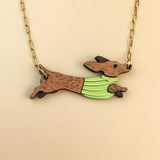 A necklace with a gold chain features a pendant of a brown Dachshund wearing a colorful sweater, depicted in a running pose against a beige background.