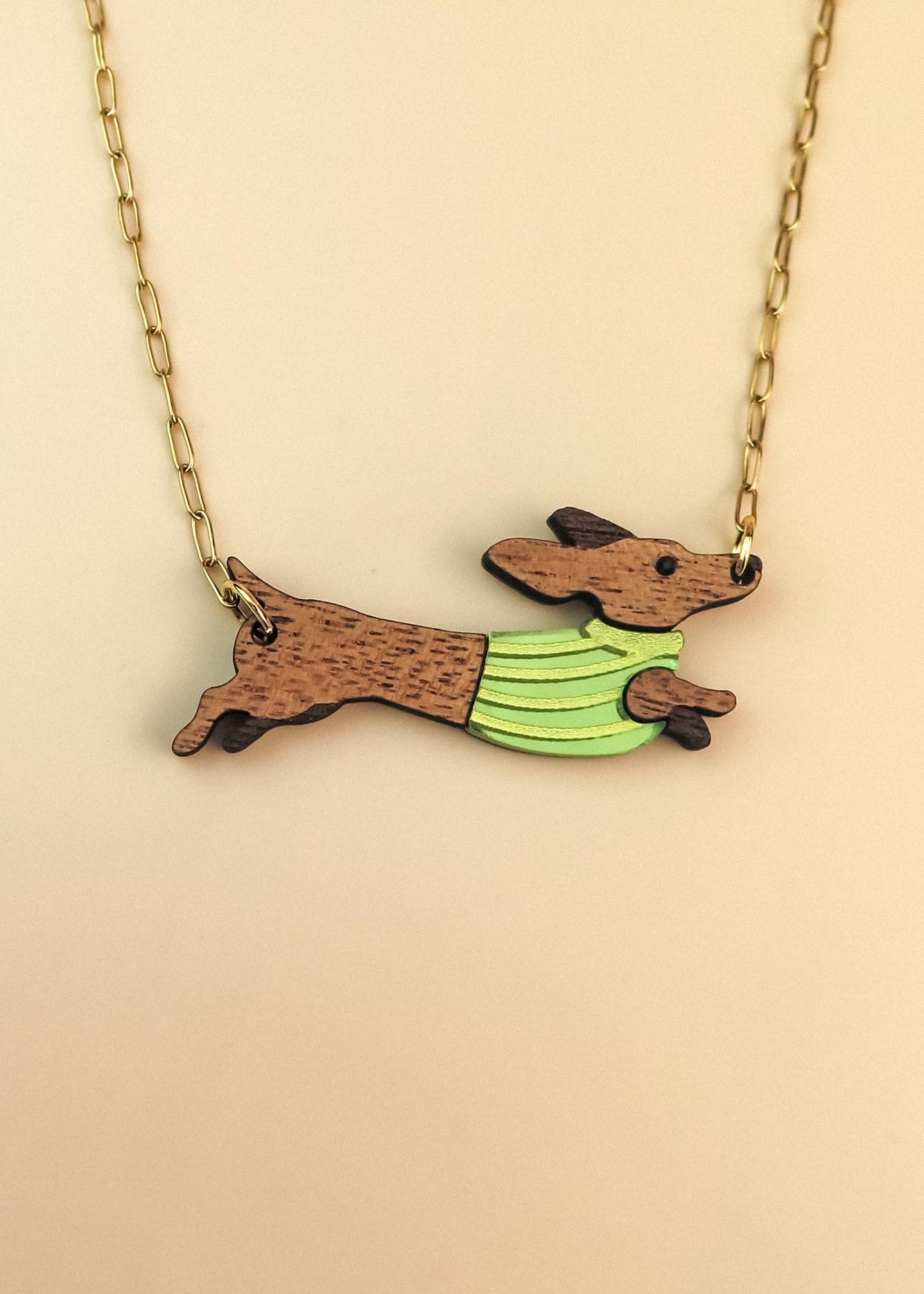 A necklace with a gold chain features a pendant of a brown Dachshund wearing a colorful sweater, depicted in a running pose against a beige background.