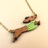 A gold chain necklace featuring a wooden Dachshund pendant in a colorful sweater, crafted from sustainable wood. The leaping pup wears green and yellow stripes against a light cream background.