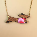 Gold chain necklace with a pendant shaped like a brown Dachshund dog, wearing a colorful sweater and crafted from sustainable wood, set against a beige background.