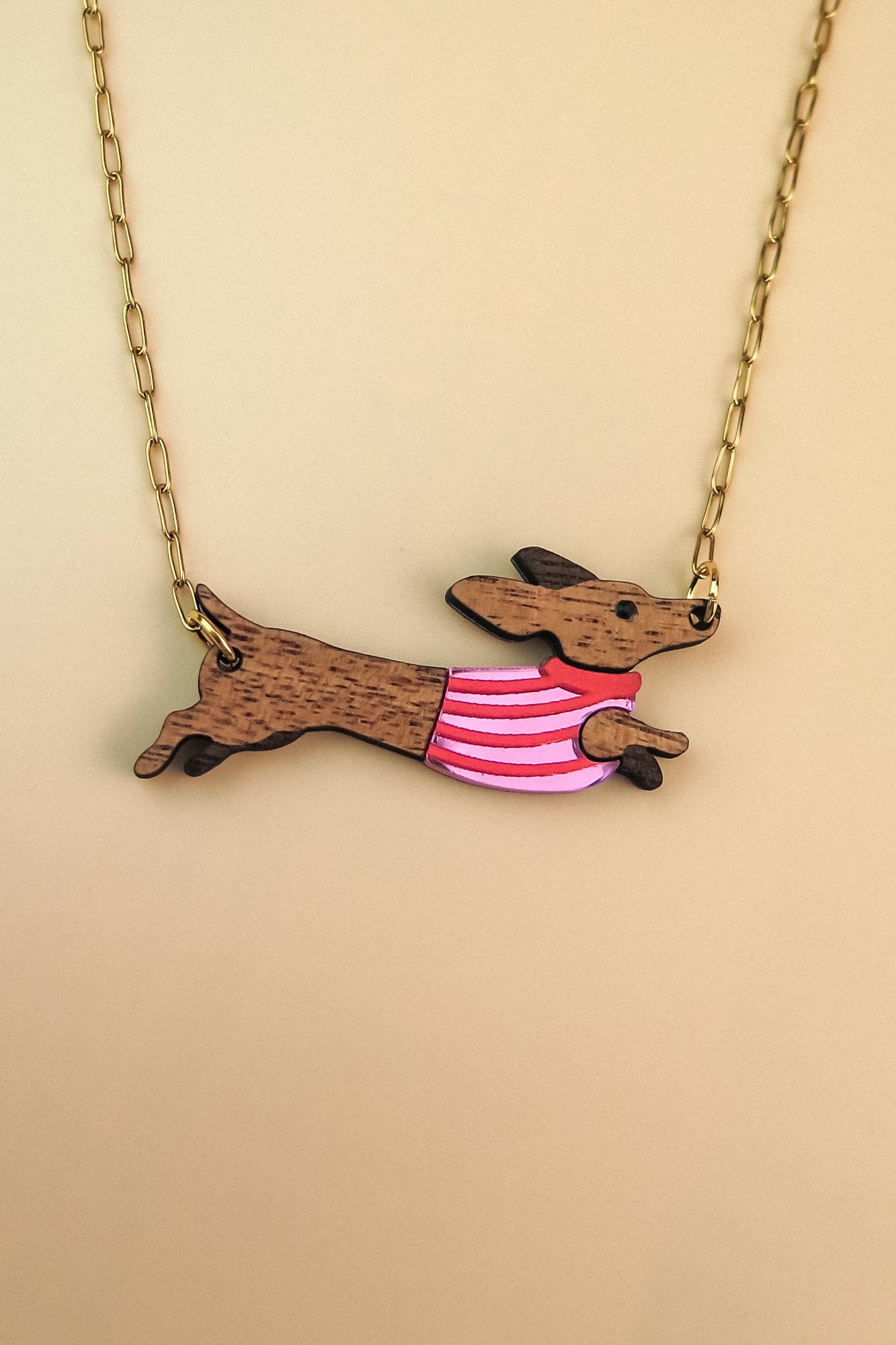 Gold chain necklace with a pendant shaped like a brown Dachshund dog, wearing a colorful sweater and crafted from sustainable wood, set against a beige background.
