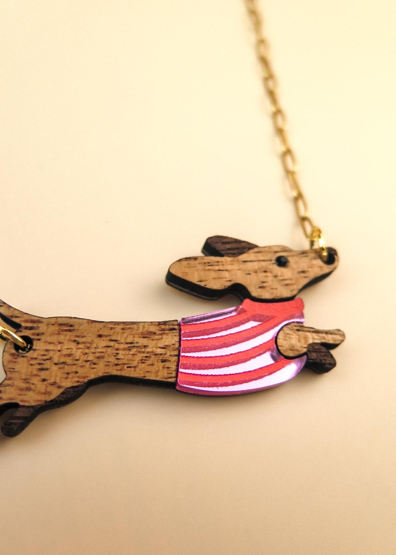 A wooden necklace pendant shaped like a dachshund dog in a colorful sweater, crafted from sustainable wood and attached to a gold chain, shown on a light beige background.