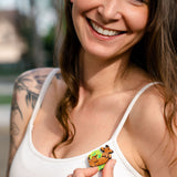 A smiling woman with long brown hair and tattoos wears a white tank top and holds a playful brooch shaped like a giraffe and leaves on her chest. The background is blurred and outdoors, highlighting her love for unique accessories like dog jewelry.