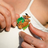 A person in a white tank top, surrounded by a green leaf backdrop, pins a playful brooch shaped like a kangaroo onto their shirt.