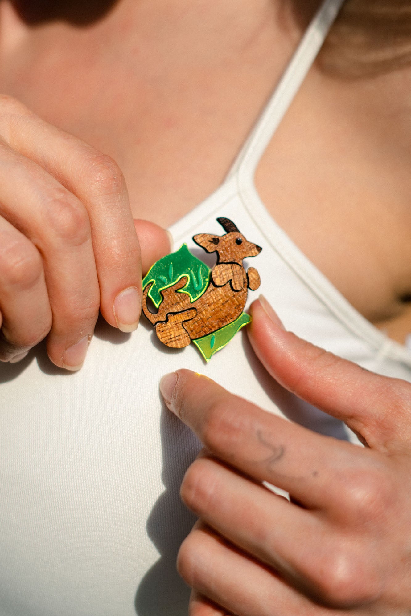 A person in a white tank top, surrounded by a green leaf backdrop, pins a playful brooch shaped like a kangaroo onto their shirt.