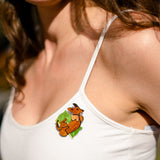 A smiling woman in a white tank top wears a playful Dachshund brooch on her chest—the colorful dog jewelry features a brown dachshund resting on a green leaf.