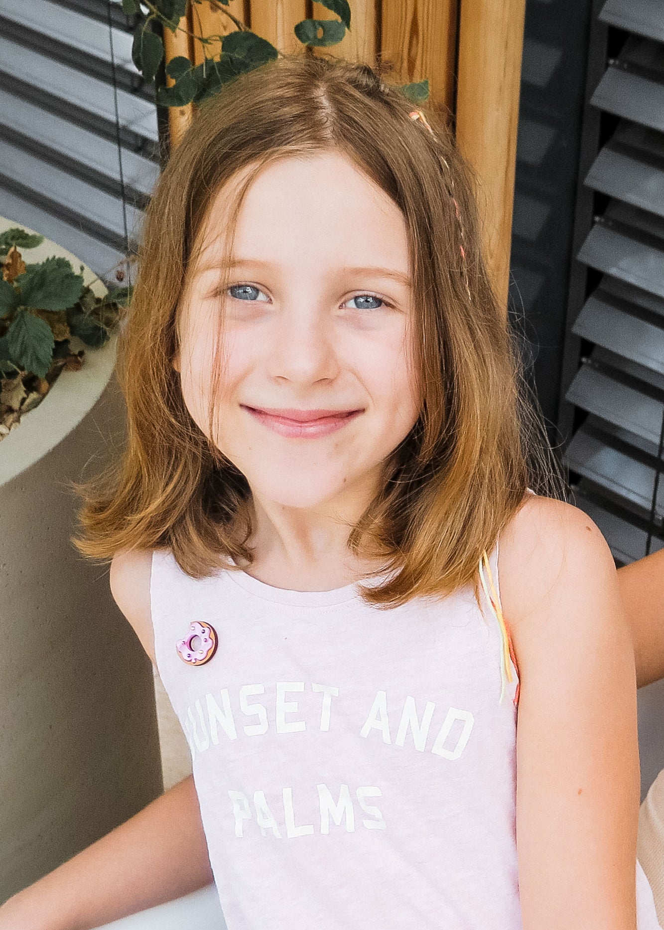 A young girl with shoulder-length light brown hair and blue eyes smiles at the camera. She wears a pale pink sleeveless top reading SUNSET AND PALMS, paired with a handcrafted Donut Pin. Plants and wooden details decorate the background.