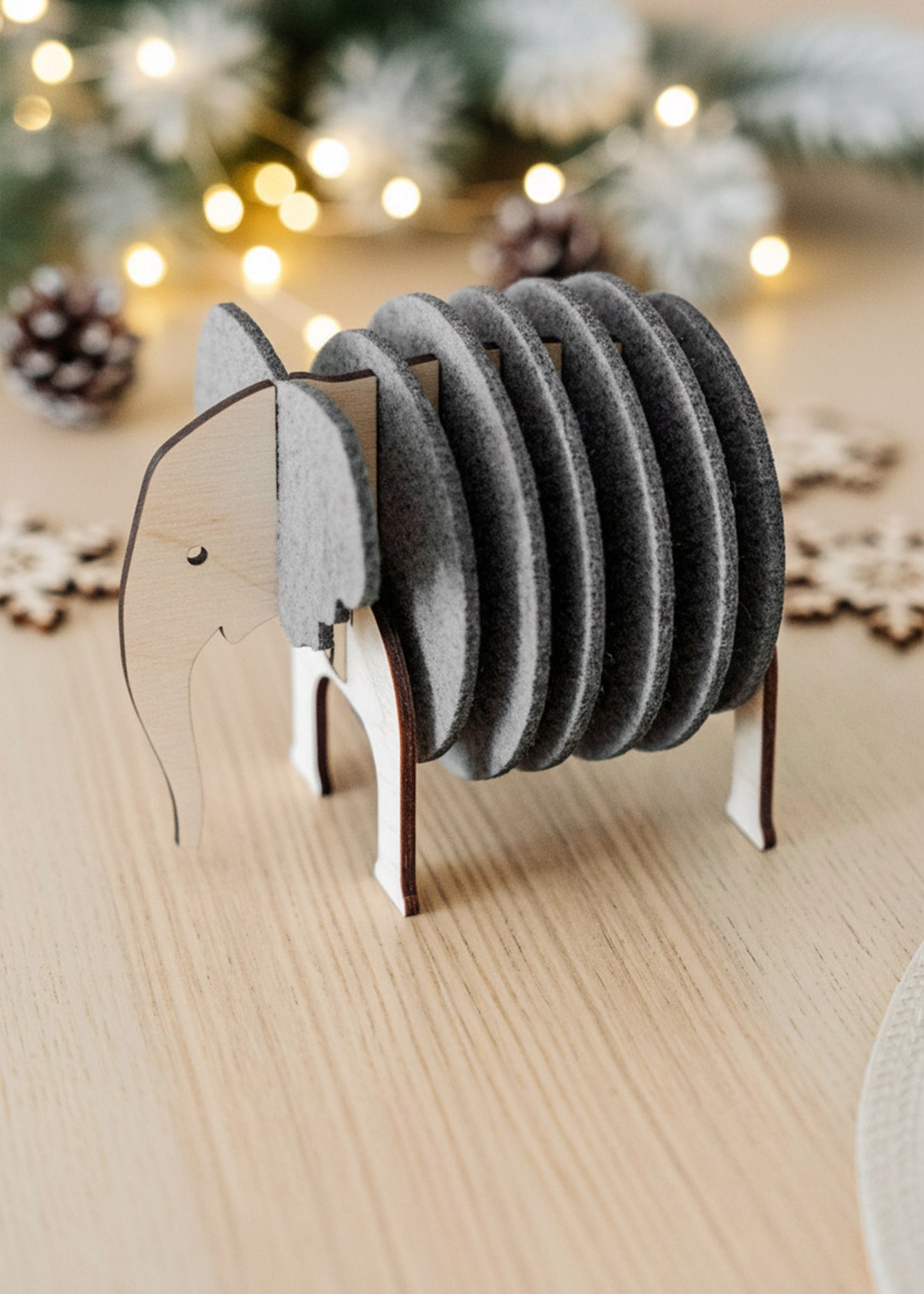 A wooden and felt elephant coasters holder sits on a light table, adding witty design and furniture protection, with pinecones, snowflake decorations, and string lights blurred in the background.