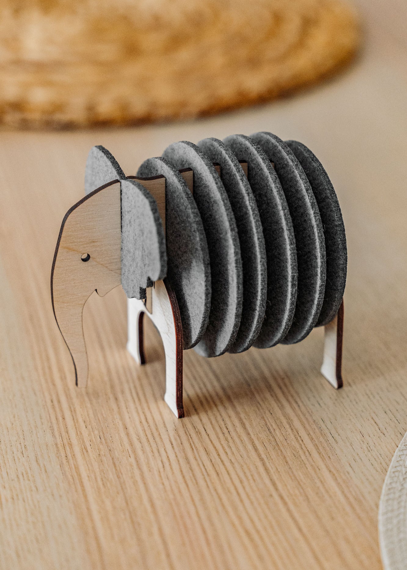 A set of round, gray Elephant Coasters is stacked vertically in a wooden holder shaped like an elephant, adding witty design and furniture protection to your light wood table.