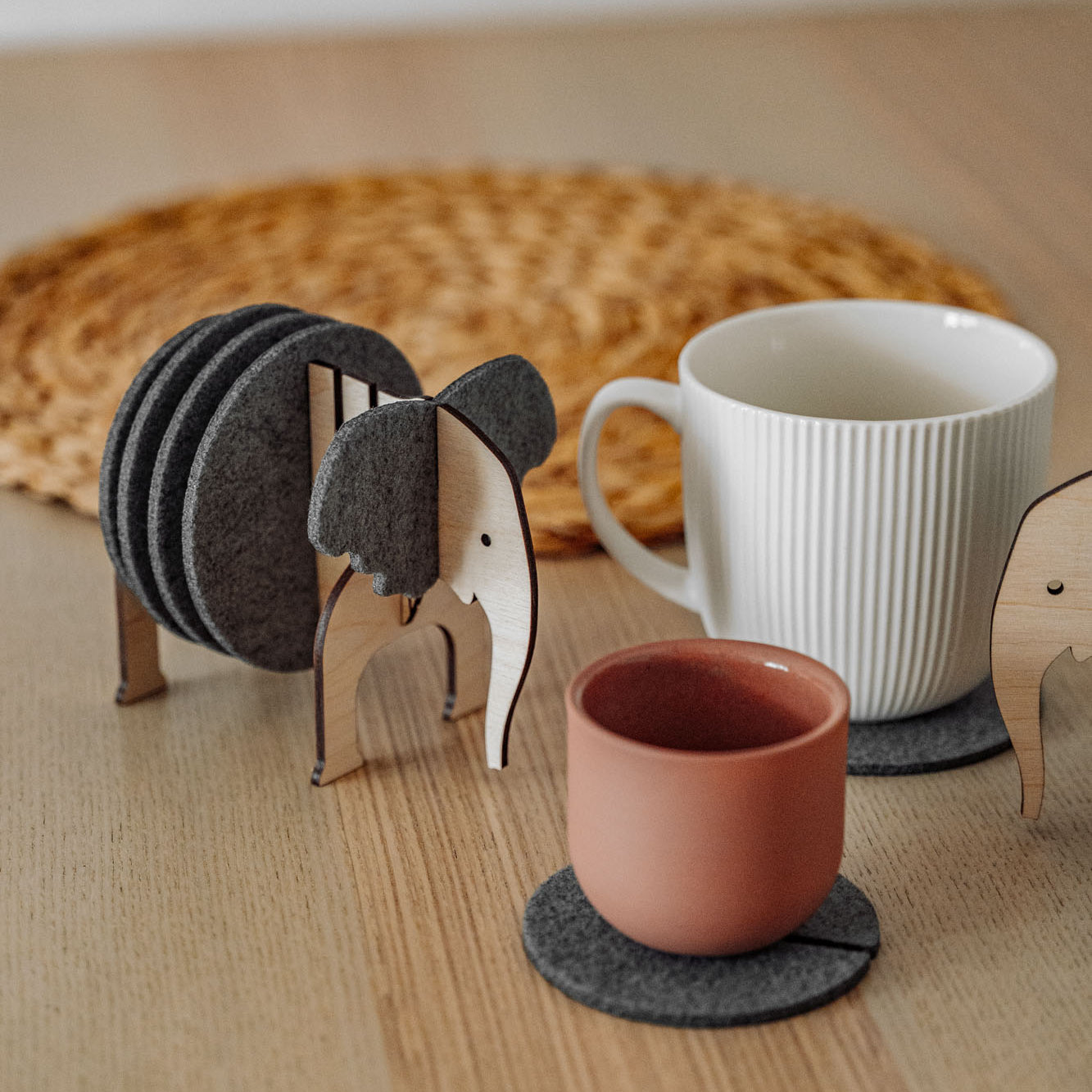 A set of Elephant Coasters with witty design offers stylish furniture protection, nestled in a wooden elephant-shaped holder on a wooden table beside a pink ceramic cup, white mug, and woven placemat.