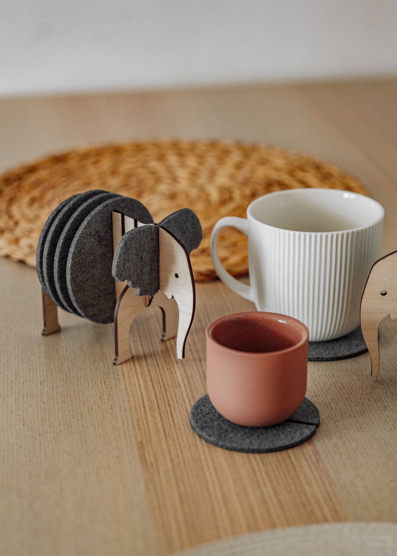 A set of Elephant Coasters with witty design offers stylish furniture protection, nestled in a wooden elephant-shaped holder on a wooden table beside a pink ceramic cup, white mug, and woven placemat.