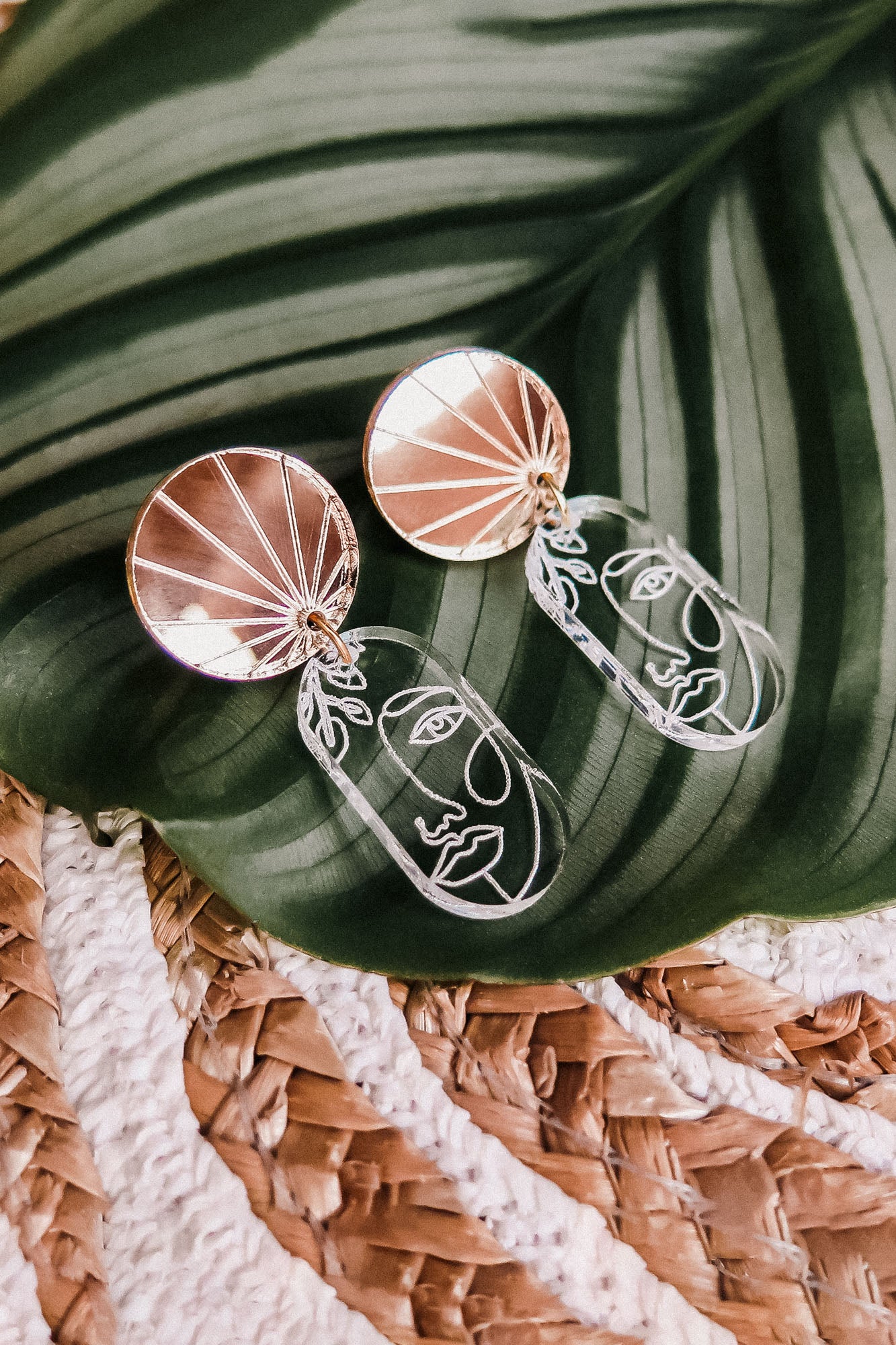 A pair of handcrafted Face Earrings with gold circular tops and abstract wire faces hang below, displayed on a green tropical leaf with a woven surface underneath to elevate your style with artful sophistication.