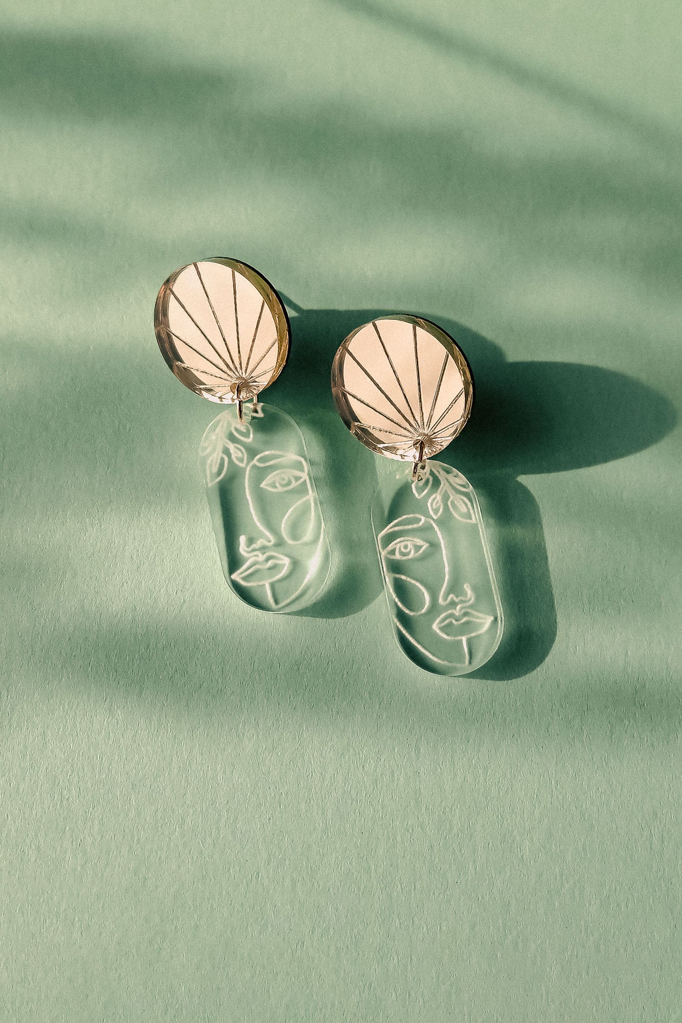 Elevate your style with these handcrafted Face Earrings: modern designs featuring round tops and dangling clear pieces etched with minimalist abstract faces, exuding artful sophistication on a green surface with soft shadows.