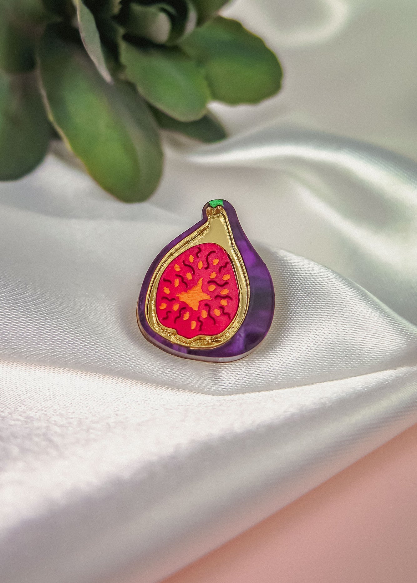 A handcrafted Fig Pin, this colorful enamel pin features a purple edge and vibrant red interior, displayed on white satin with green leaves behind. A lightweight accessory that adds a pop of color to any look.