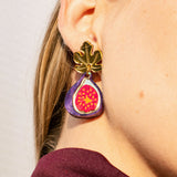 Close-up of a woman’s ear wearing handcrafted Fig Earrings shaped like a gold fig leaf with a colorful fig fruit design hanging below it. The woman has light skin, straight blonde hair, and the delicate earrings shimmer like gold mirror leaves.