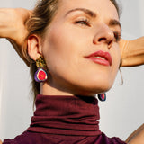 Woman wearing a sleeveless maroon turtleneck top and handcrafted fig-shaped earrings with delicate gold mirror leaves poses with arms raised behind her head, looking upward against a light background.