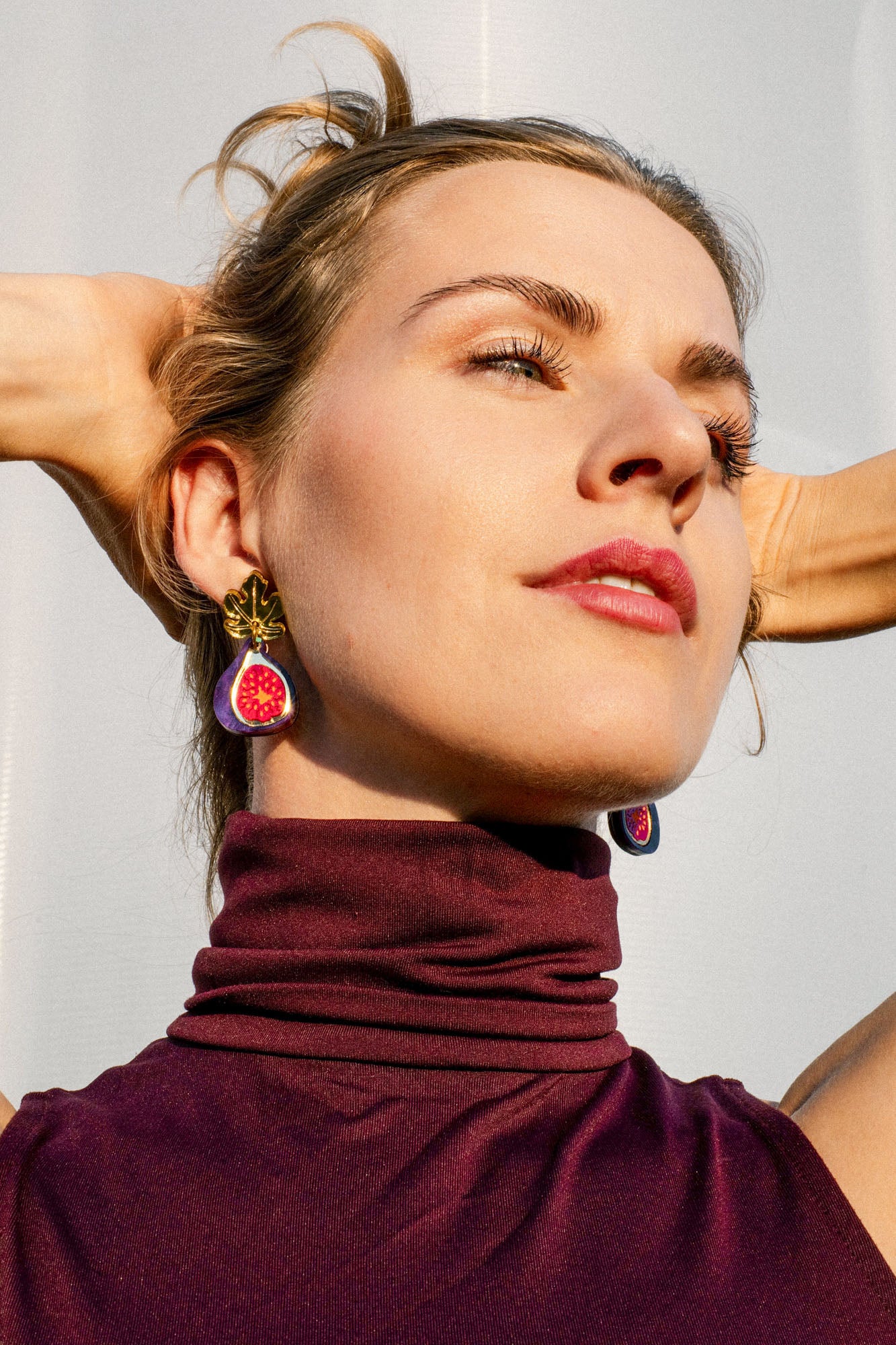 Woman wearing a sleeveless maroon turtleneck top and handcrafted fig-shaped earrings with delicate gold mirror leaves poses with arms raised behind her head, looking upward against a light background.