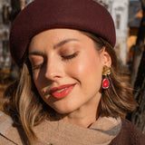 A woman outdoors smiles gently with her eyes closed. She wears a burgundy beret, a tan scarf with a fig brooch, and handcrafted fig earrings accented with gold mirror leaves. Autumn trees and buildings are blurred in the background.
