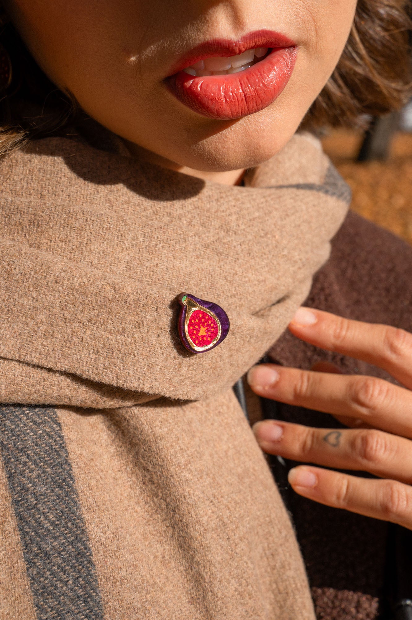 A person wearing red lipstick and a beige scarf with a handcrafted Fig Pin, a unique accessory shaped like a fig; their hand rests on the scarf, showing neat nails and a small finger tattoo.