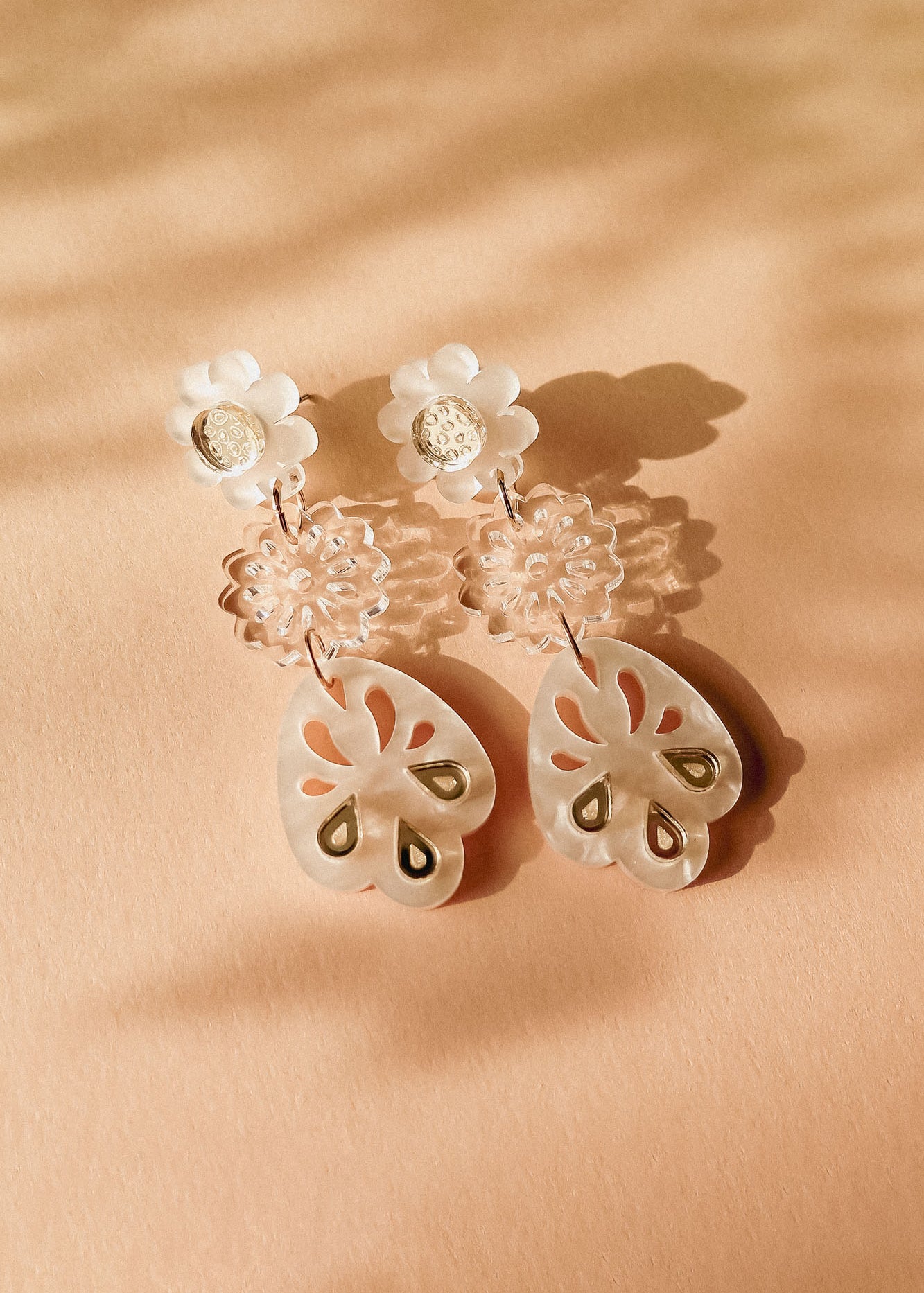 A pair of handcrafted dangling earrings with white floral studs, clear floral middle segments, and translucent, petal-shaped drops featuring cut-out and gold accents, displayed on a peach-colored surface with soft shadows.