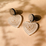 A pair of Folk Heart Earrings with wooden circular studs and intricate, lace-like craftsmanship form lightweight elegance, resting on a beige surface with soft shadows.