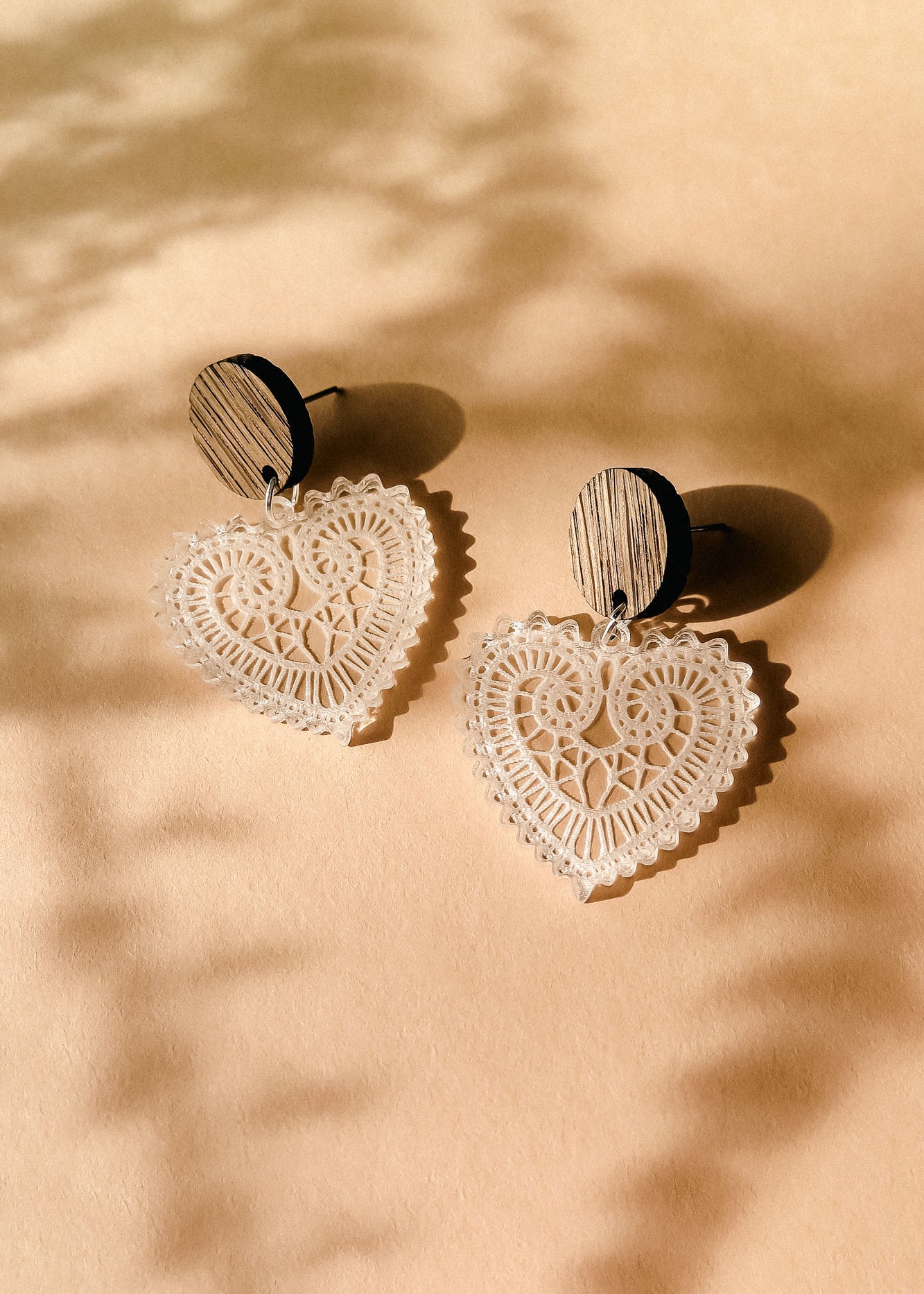 A pair of Folk Heart Earrings with wooden circular studs and intricate, lace-like craftsmanship form lightweight elegance, resting on a beige surface with soft shadows.